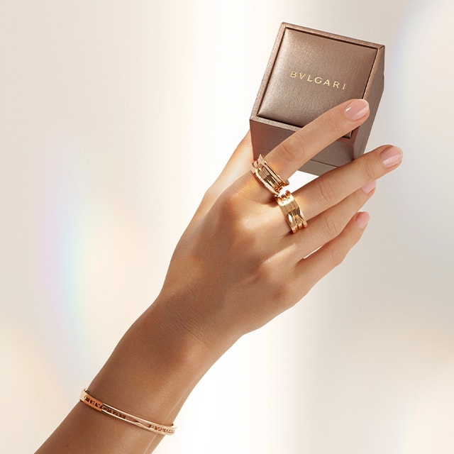 Hand with B.zero1 rings and bracelets holding a Bulgari gift box and B.zero1 four-band rose gold ring.