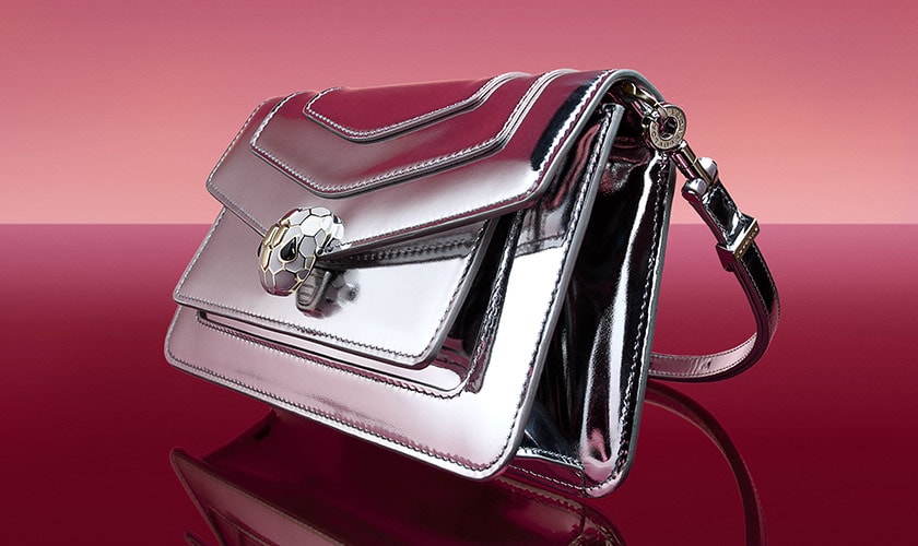 Serpenti Forever bag in silver mirror calf leather with snakehead closure, dark red backdrop.