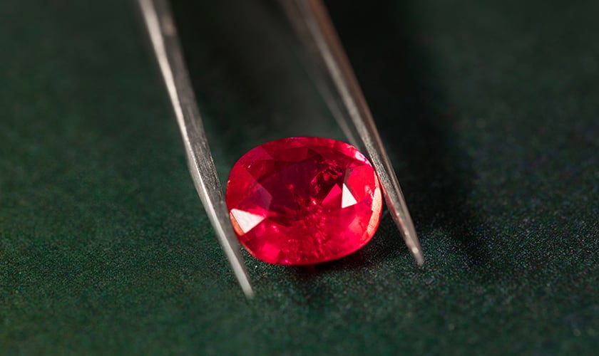 A Bulgari cushion-cut ruby held by tweezers, dark background, close up.
