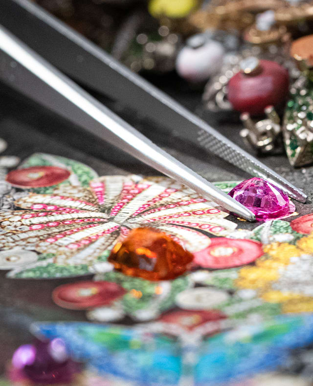 Craftsman placing a coloured gem with a tweezer on the sketch of a Bulgari necklace with flowers, close up.