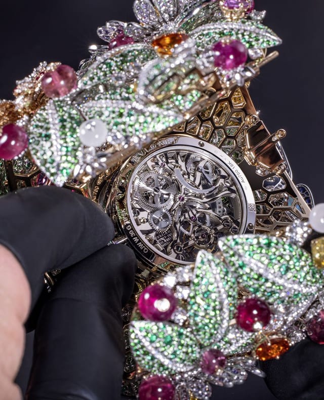 Hands with black gloves holding a High Jewellery watch in the making set with coloured gemstones.