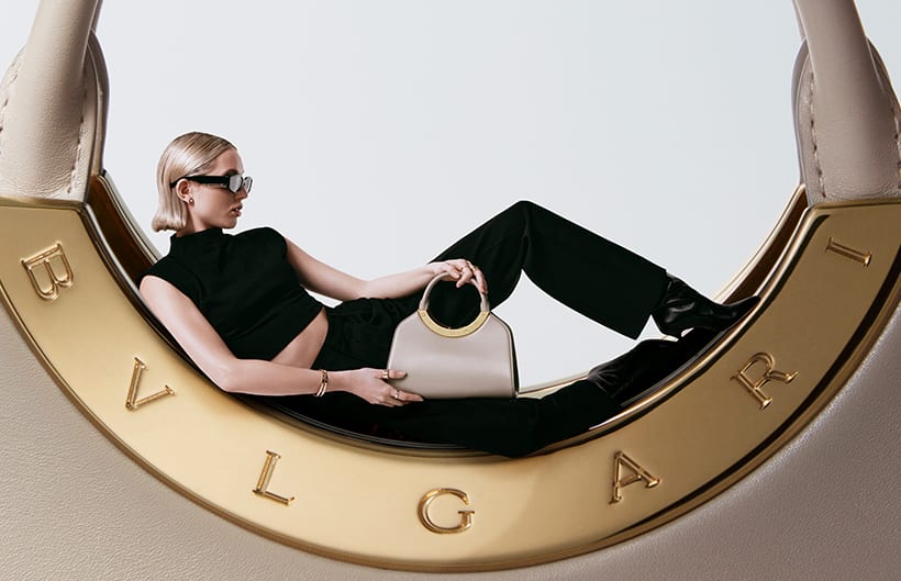 Model lying on the half-moon metal detail with Bulgari logo of a giant Bvlgari Roma bag in ivory calf leather.