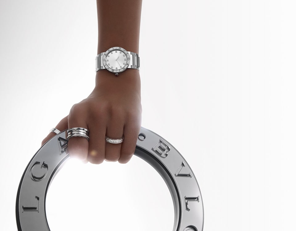 Hand wearing a Bvlgari Bvlgari steel watch and holding a metallic circle with a Bvlgari Bvlgari engraving.