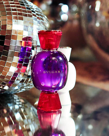 Partying model holding the Bvlgari Fantasia Veneta fragrance bottle in purple and red glass and the white Magnifying Essence flacon.