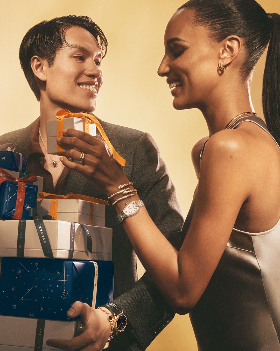Woman unwrapping a Bvlgari gift box, adorned with jewels beside a man holding stacked presents.